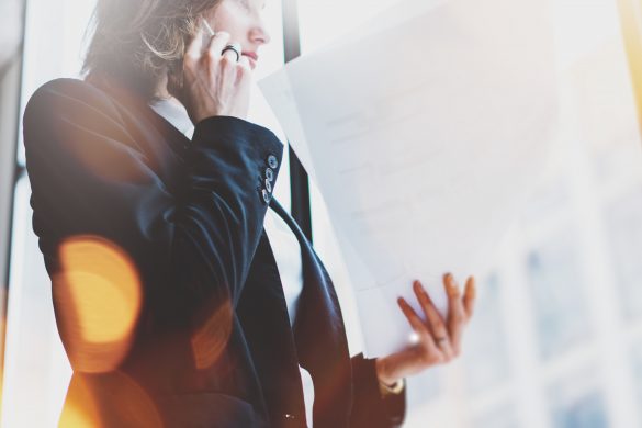 Photo business woman wearing white shirt, talking smartphone and holding documents in hands. Open space loft office. Panoramic windows background. Wide mockup